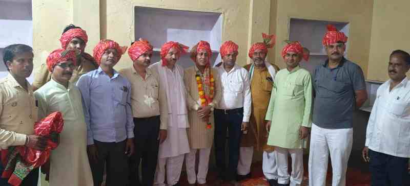 कई पुरुष पारंपरिक राजस्थानी साफा और माला पहनकर दीवार के सामने समूह में खड़े हैं