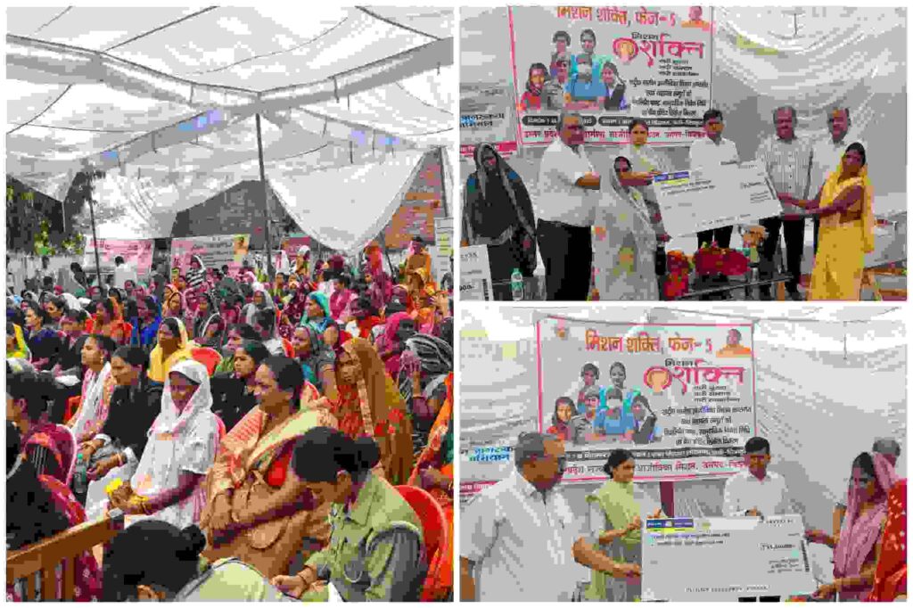 "महिलाओं की भीड़ और मंच पर प्रतिनिधियों द्वारा बड़े चेक वितरित करते हुए मिशन शक्ति योजना का आयोजन"
