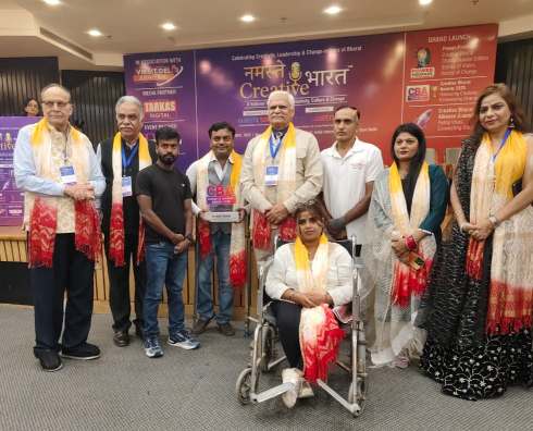 भारत के एक सम्मान समारोह में विभिन्न प्रतिभागी पारंपरिक वस्त्र और सम्मान पट्टिका पहनकर मंच पर खड़े हैं, एक प्रतिभागी व्हीलचेयर पर है