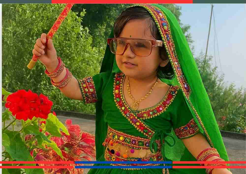 "हरी रंग की पारंपरिक गरबा ड्रेस पहने नन्ही बालिका हाथ में डांडिया स्टिक लेकर गरबा की मुद्रा में, बैकग्राउंड में रंग-बिरंगे पौधे।"