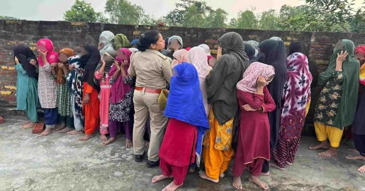 पुलिस अधिकारी की अगुवाई में महिलाओं की लंबी कतार, सभी ने अपने चेहरे को कपड़ों से ढक रखा है और दीवार के सामने खड़ी हैं।