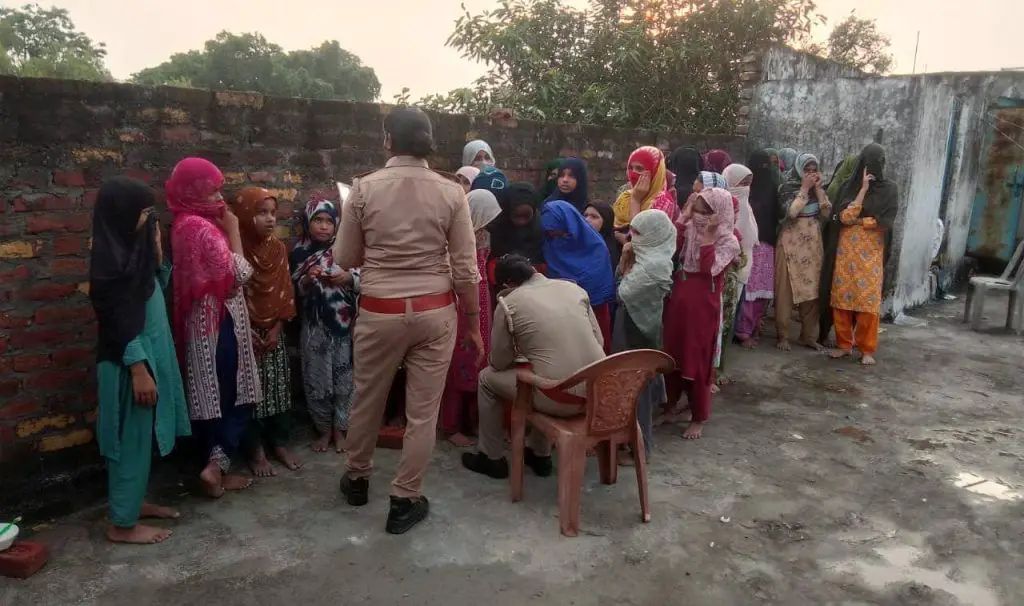 एक दीवार के सामने खड़ी महिलाओं की भीड़ को दो पुलिस अधिकारी सहायता देते हुए, महिलाएं अपने चेहरे दुपट्टे से ढक रही हैं।