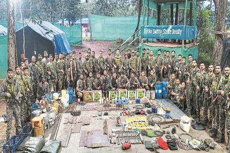 फौजी वर्दी में दर्जनों कमांडो जवान, जब्त किए हुए हथियार, हथगोले, गोलियां, विस्फोटक और अन्य सामग्री के साथ समूह में खड़े हैं; पास में कैंप और एक चौकी भी दिख रही है
