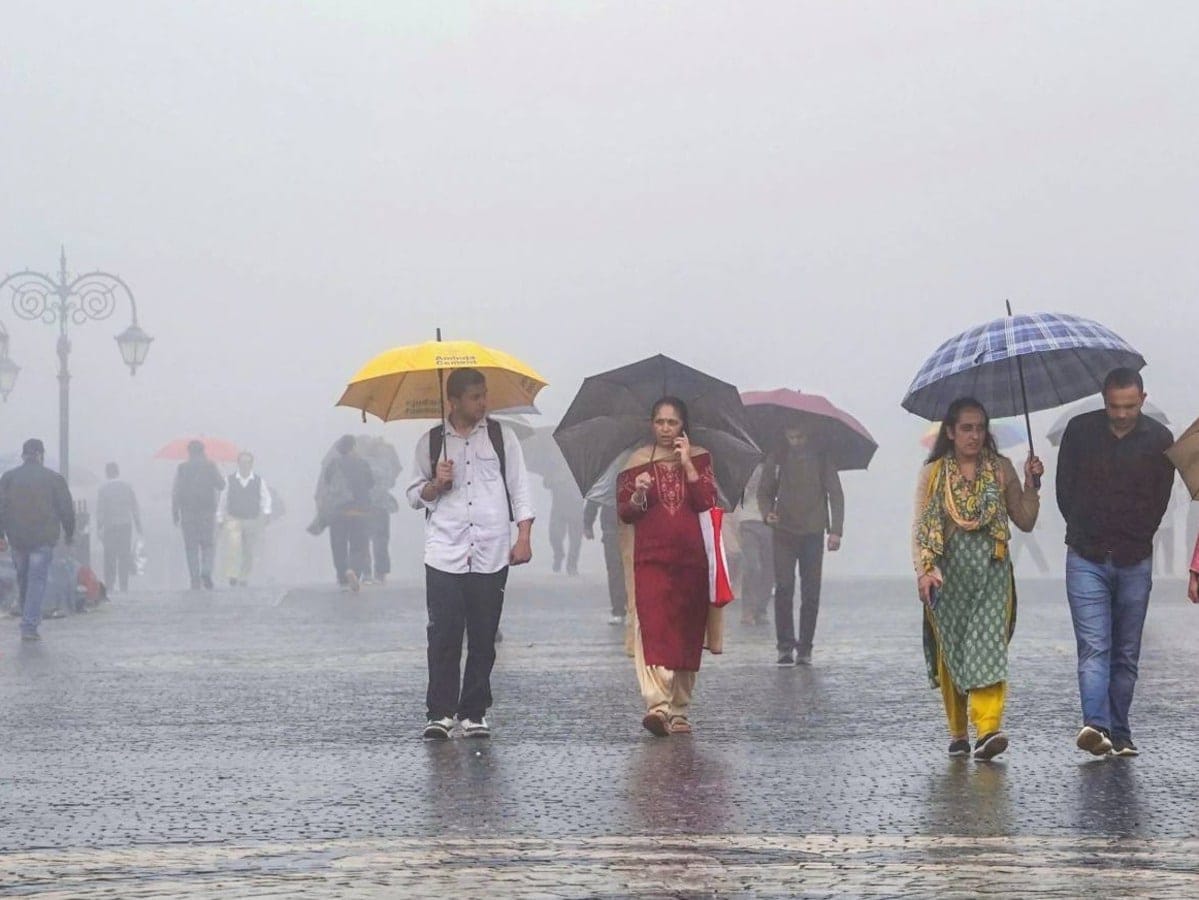 दिल्ली की सड़कों पर बारिश के दौरान लोग विभिन्न रंग के छाते लिए चल रहे हैं, धुंध और नम वातावरण दिखाई दे रहा है।
