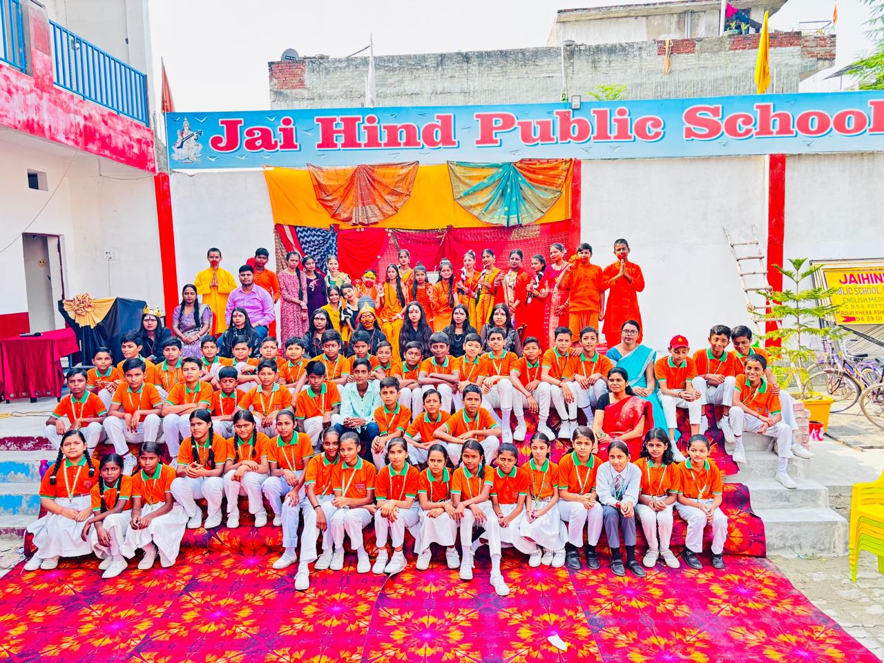 अशरफ नगर स्थित जय हिंद पब्लिक स्कूल के छात्र और शिक्षक दीपावली उत्सव 2025 के अवसर पर समूह फोटो में, छात्र नारंगी शर्ट और सफेद पैंट/स्कर्ट में, शिक्षक पारंपरिक पोशाक में, रंग-बिरंगे मंच और सजावट के सामने खड़े हैं।