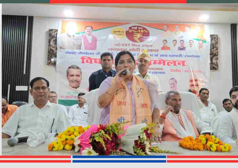 सम्मेलन में मंच पर महिला वक्ता माइक के साथ, बैकग्राउंड में बैनर और बैठे हुए कई लोग