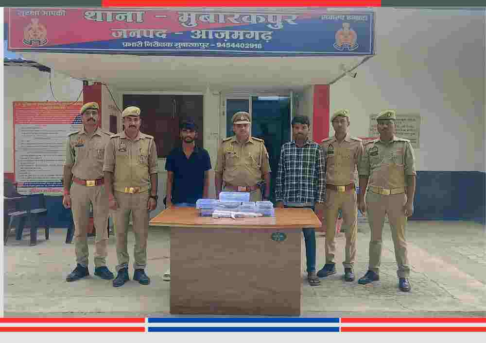 छह पुलिसकर्मी और दो गिरफ्तार व्यक्ति एक टेबल के पीछे खड़े हैं, टेबल पर जब्त सामान रखा है, पीछे थाना मुबारकपुर, जनपद आजमगढ़ की नेम प्लेट लगी है।
