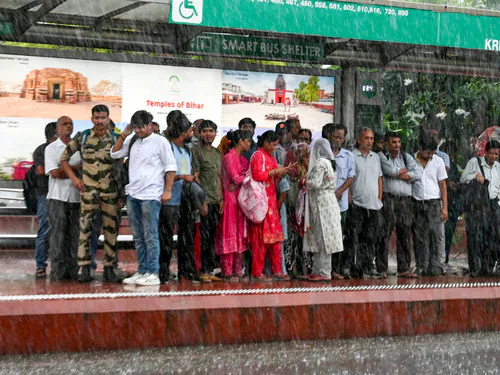दिल्ली-एनसीआर में तेज बारिश के दौरान बस स्टॉप पर खड़े लोग, कुछ छाता लिए हुए हैं, जबकि कई भीग रहे हैं।