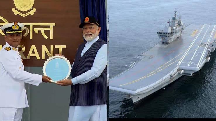 भारतीय नौसेना का अधिकारी और एक गणमान्य व्यक्ति मंच पर सम्मान समारोह में प्लेट थामे हुए, साथ ही समुद्र में खड़ा भारतीय विमानवाहक पोत।