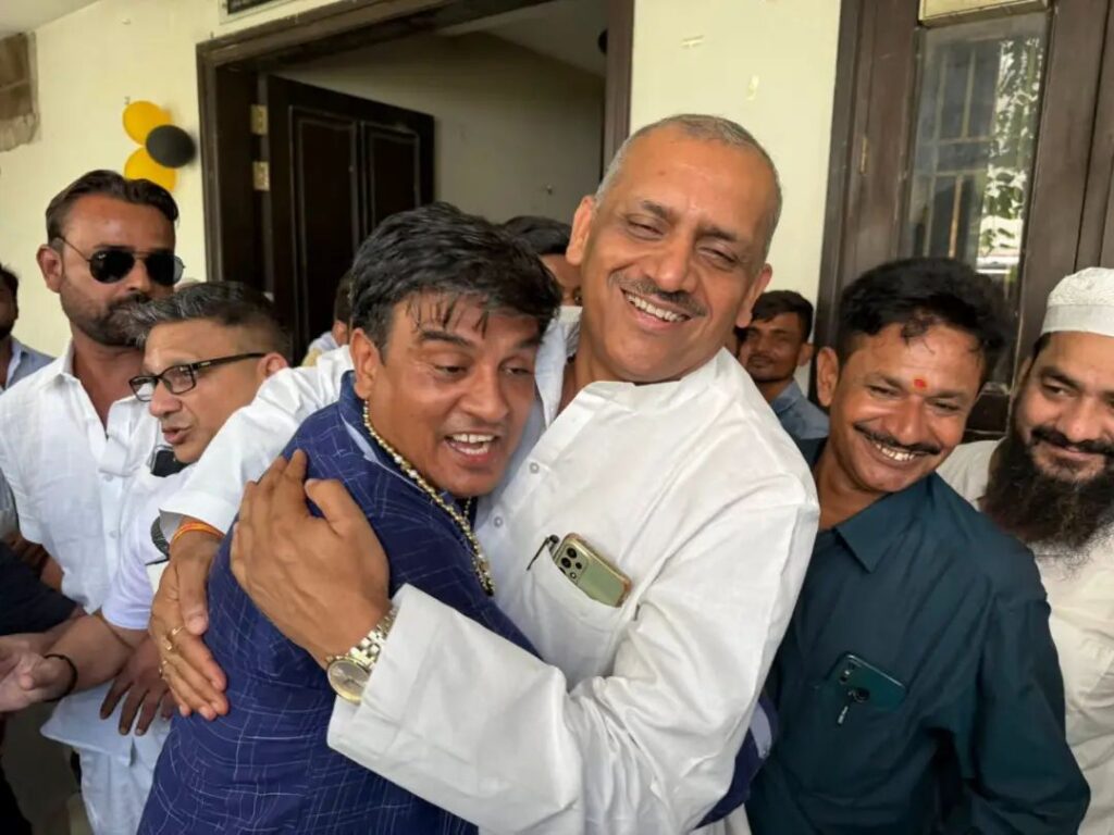 समारोह में दो व्यक्ति गले मिल रहे हैं, उनके आस-पास कई लोग मौजूद हैं